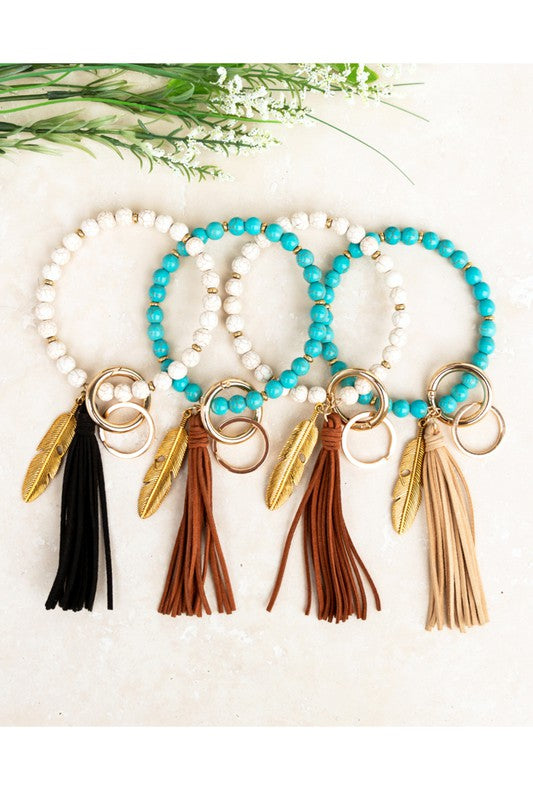 Boho Stone Key Ring Bracelet Earthy, boho vibe featuring genuine stone beads, suede tassel, and feather charm. Key Ring Bracelet Genuine Stone Antique gold feather charm Removable Gold Clasp Removable Gold Key Ring Reinforced Wire design Dimensions: 4" Suede Tassel Gold Accent Beads